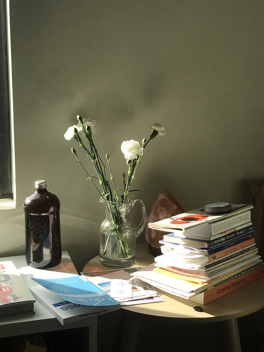 a stack of books, a jug with water and flowers, a salt lamp, on a bedside table in the sun