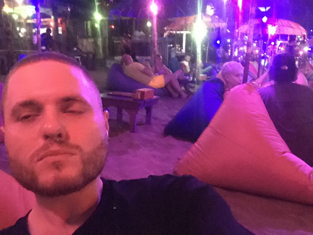 Me (Louis) half blinking while I try to take a selfie on the beach, at a beach bar with bean bags and purple lights