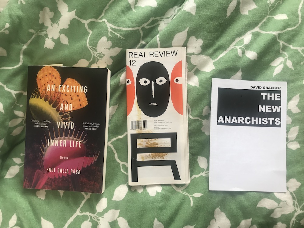 a green bedspread with these three books ontop: The New Anarchists by David Graeber, Real Review issue 12, An Exciting and Vivid Inner Life by Paul Dalla Rosa