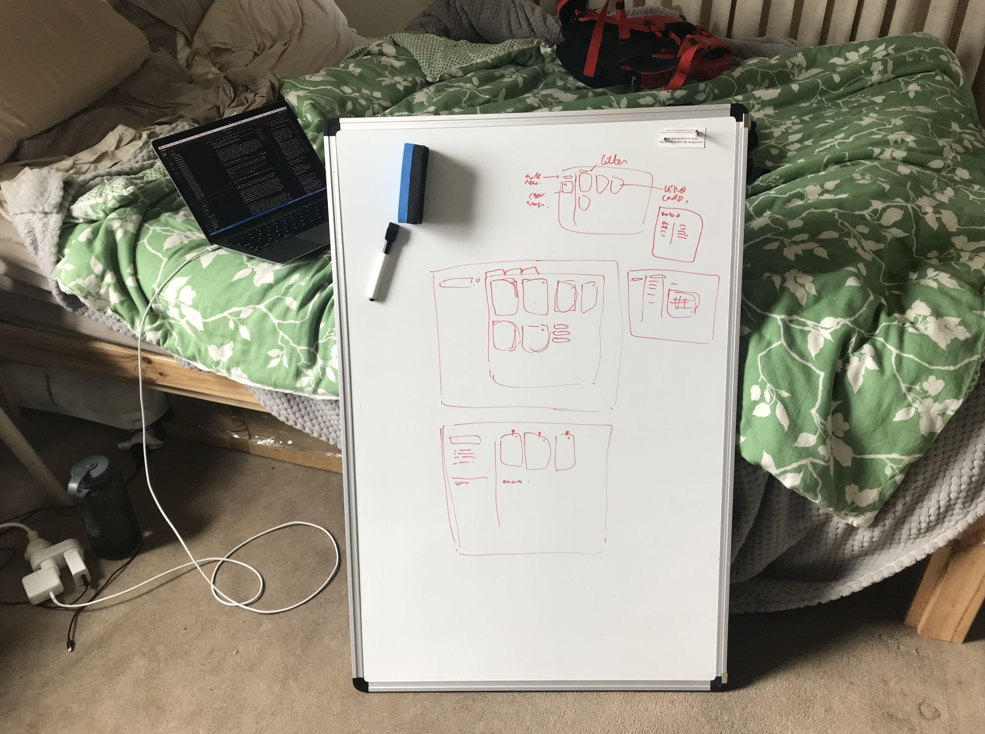 a whiteboard with wireframes drawn on it, leaning on a messy bed with a laptop behind it