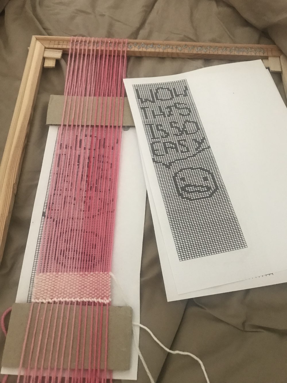 a loom on the bed with a tapestry started on it. There is a printed design behind the warp and a copy beside it, with a small face and text that reads "wow this is so easy"