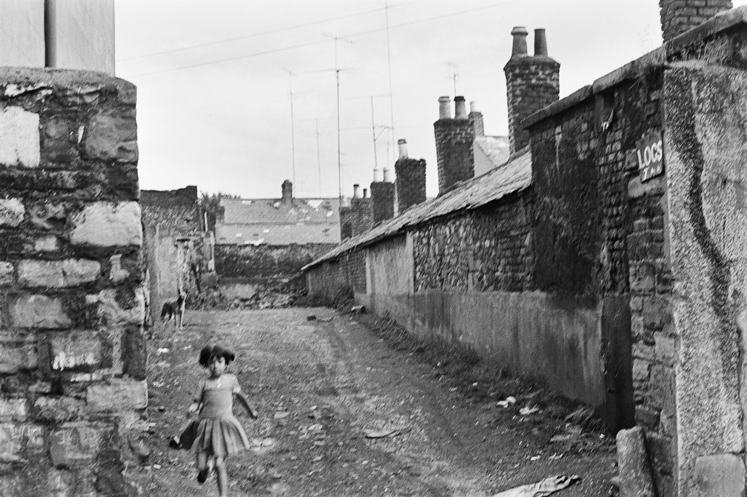 a blurry black and white photo of a wide backalley, a child is running out of it and a dog is in the background